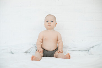 Beautiful little blue eyed baby sit on white sheet on bed and look up, free copy space. Portrait of charming naked infant child on white background. Concept of happy childhood and childcare