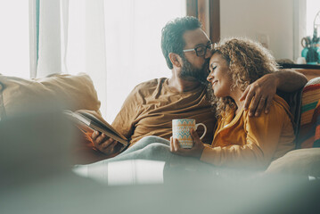 Couple of young adult man and woman enjoy time together sitting on the sofa in indoor leisure activity.. Real life people at home. Male and female relax on the couch reading a book and drinking a tea - Powered by Adobe