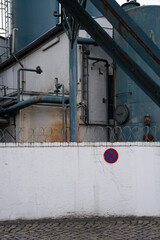 Industrial piping of a chemical plant. Painted in turquoise. Rusty steel construction, barbed wire and a white painted wall with traffic sign "no parking", "parking prohibited". Copy space available.