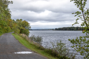 Fototapeta na wymiar Gravel bicycle path along a river under dark cloudy skies, forest and grass along the path, nobody
