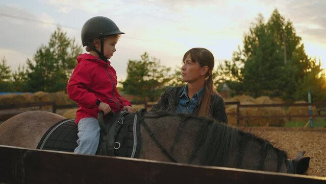 therapeutic riding instructor woman is talking with little boy on horseback in equestrian club during training