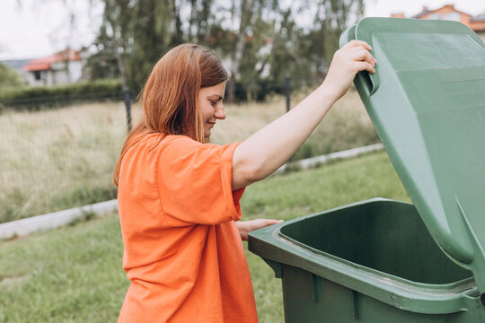 Young happy woman putting garbage in green trash bin on back yard background. Environmental conservation saving planet from contamination. Plastic pollution problem