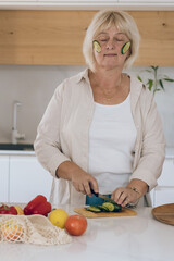 Middle Age Woman Cutting Vegetable On Kitchen Board And Undergoing A Cucumber Facial Treatment.