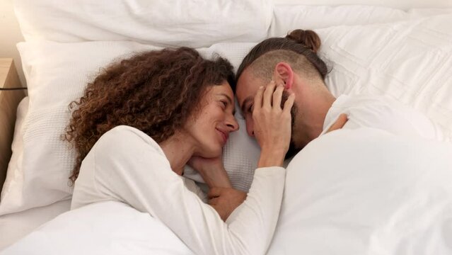 Couple in bed love bonding, smile and laughing at funny comedy, comic or goofy joke together in house. Silly, romance and happy young man and woman touch face, loving and wake up in apartment bedroom