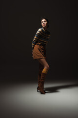 full length of young woman in turtleneck with skirt and boots posing on black.