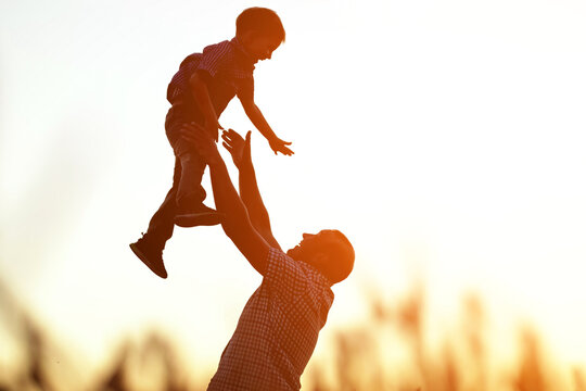 Father Silhouette Tosses Preschooler Boy On Wheat Field. Son Enjoys Holidays And Looks With Excited Expression Against Clean Sky In Countryside, Sunlight