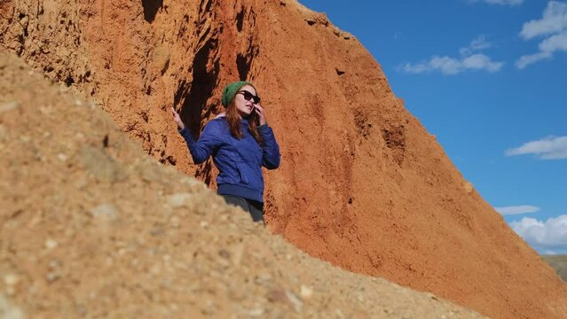 Excited young woman standing among red rocks on blue sky background, looking around. Female hiker admires beautiful mountains.