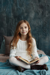 Young pretty girl with long curly hair dressed in white knitted sweater, boots sitting on the bed with blanket and pillow, reading a book, looking at the camera and smiling, cozy warm winter portrait