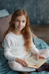 Young pretty girl with long curly hair dressed in white knitted sweater, boots sitting on the bed with blanket and pillow, reading a book, looking dreamy at camera, cozy warm winter portrait