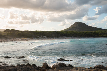 The waves splash on beach of Cala Agulla (Cala Ratjada) on Mallorca in the mediterranean sea. Blue water and a green mountain in the background. People bathing, surfers surfing. Tropical paradise.