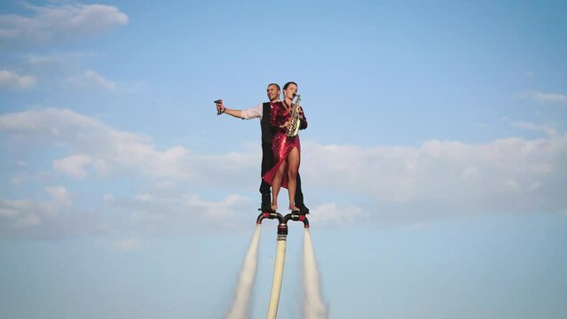 Show over the water on a flyboard. A woman in a red dress plays the saxophone, a man flies with her together.