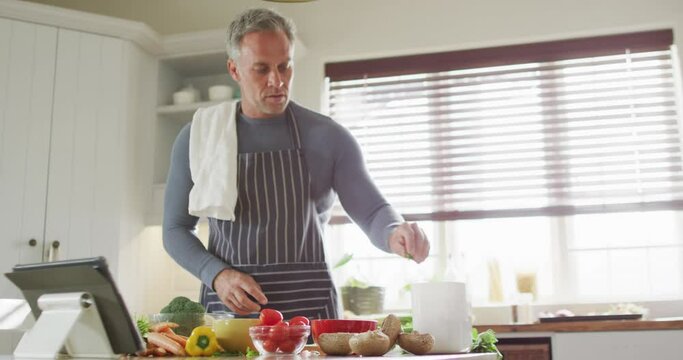 Happy caucasian man wearing apron, standing in kitchen, cooking dinner and using tablet