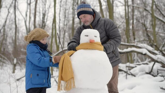 Little boy with his father building snowman in snowy park. Dad and son spend quality time together. Active outdoors leisure with family with children in winter. Kid during stroll in a snowy winter par