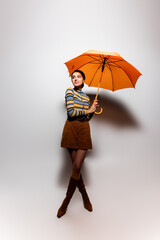 full length of smiling young woman in striped turtleneck and skirt posing with orange umbrella on grey.
