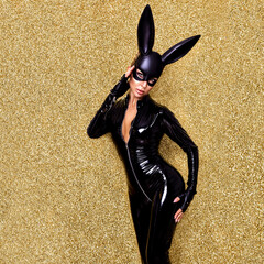 Sexy blonde woman posing in Halloween vinyl black costume and black bunny mask on on a glitter gold...
