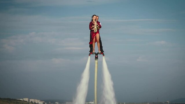 Show over the water on a flyboard. A woman in a red dress plays the saxophone, a man flies with her together.