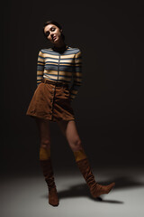 full length of young woman in turtleneck with skirt and autumnal boots posing on black.