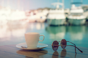 White cup with saucer, sunglasses on a table on the background of blurred boat harbor. Summer vacation concept