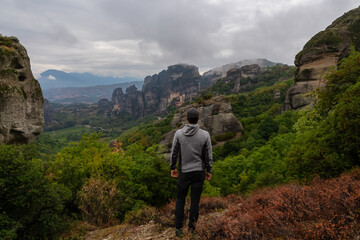Fototapeta premium Man with scenic view of Holy Monastery of Rousanos and Holy Monastery St Nicholas Anapafsas appearing from fog, Kalambaka, Meteora, Thessaly, Greece, Europe. Misty atmosphere in dramatic landscape