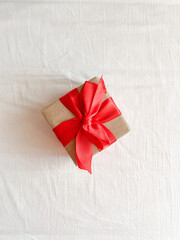 The gift, tied with a red ribbon and wrapped in kraft paper, is on a white textile surface. Copy space. Top view. Flatlay