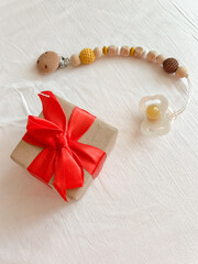 Wooden beads-holders with a pacifier and a gift with a red ribbon are on a white smooth textile surface. Christmas gift for a newborn