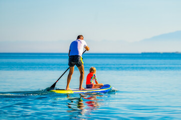 Father and daughter riding SUP stand up paddle on vacation. - obrazy, fototapety, plakaty