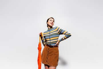 pleased woman in striped turtleneck and skirt posing with hand on hip and holding orange umbrella on grey.