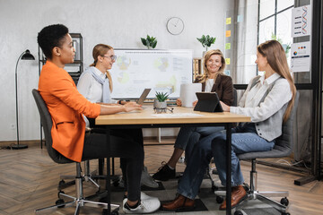 Attractive businesswomen sitting together at modern office and working on tablets and laptops. Competent brokers analyzing charts on trade market.