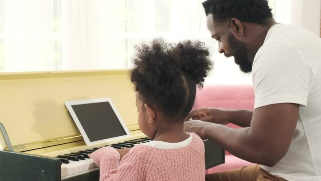 Black girl and father play piano together and using tablet for reading music note in living room.