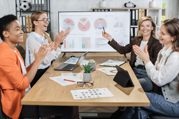 Multi ethnic female partners in formal clothes applauding together to their mature colleague while sitting at office. Competent financiers with modern gadgets on table looking and smiling on camera.