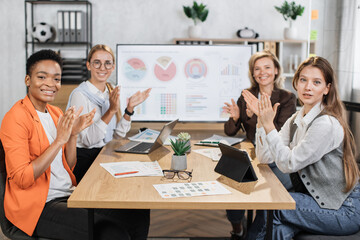 Multi ethnic female partners in formal clothes applauding together while sitting at office. Competent financiers businesswomen with modern gadgets on table looking and smiling on camera.