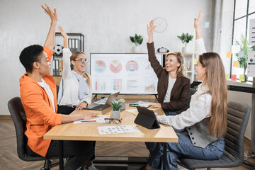Team of multi ethnic female financial experts celebrating successful negotiation while sitting at desk and holding hands up. Women using modern gadgets for work at office.