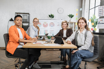 Female economists in formal wear looking at camera while sitting together at office desk with modern gadgets and documents. Multi ethnic women brokers spending evening for examining trade market.