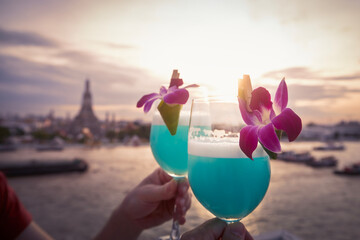 Fototapeta premium Cocktail drinks with blue curacao. Couple holding decorated drinking glasses against city view at sunset. Bangkok, Thailand.