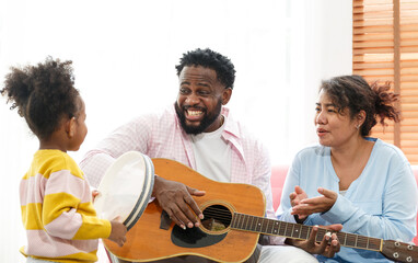 Cute little girl with mom and dad playing guitar and smiling while sitting on the sofa at home. singing fun music