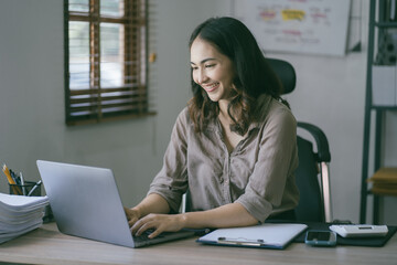 Portrait of a beautiful Asian woman looking at laptop screen while sitting at working desk in the office