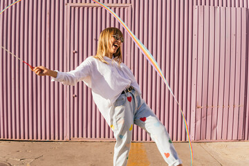 Cheerful woman playing with rainbow colored gymnastics ribbon in front of corrugated wall