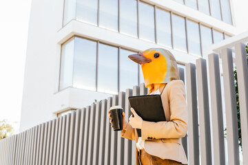 Freelancer wearing bird mask standing with coffee cup and tablet PC in front of building