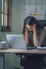 Portrait of a beautiful Asian woman looking at laptop screen while sitting at working desk in the office