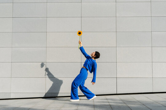 Woman with sunflower bending over backwards in front of wall on sunny day