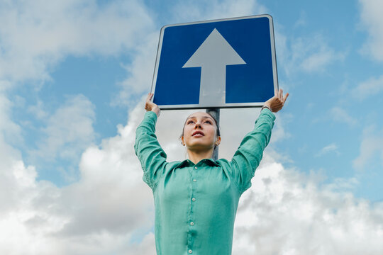Businesswoman with arrow sign board standing under cloudy sky