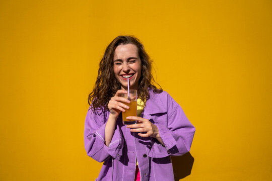 Cheerful woman with eyes closed drinking juice in front of yellow wall