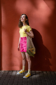 Smiling woman carrying mesh bag day dreaming in front of red wall