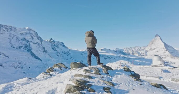 Aerial orbit motion of Young asian man standing on top of the famous Gornergrat observatory operates with drone remote control with iconic famous Matterhorn sunset background. Swiss alps, Switzerland.