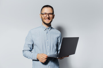 Studio portrait of young smiling man holding a laptop in hands, wearing blue shirt and eyeglasses on white background.