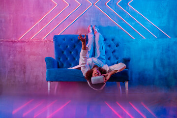 Smiling woman with futuristic glasses lying upside down on sofa in front of neon light