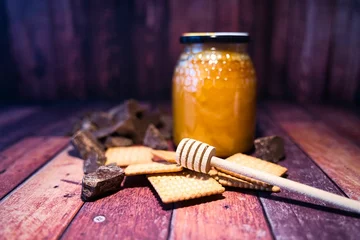 Tuinposter Jar of bee honey with a honey dipper, some cookies, and dark chocolate placed on a wooden surface © Saltacekias/Wirestock Creators