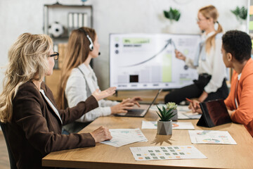 Group of competent diverse female economists discussing business ideas while gathering at office room. Caucasian blond woman standing near big monitor with various graphs and charts.