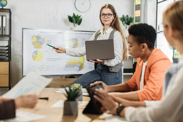 Caucasian woman presenting financial report on monitor for multiracial female colleagues at modern office. Business women partners sitting at desk. Business conference concept.