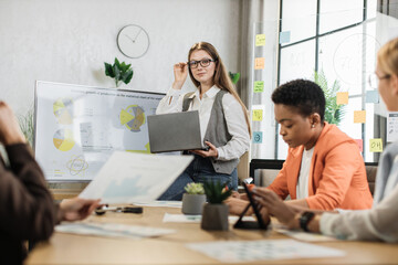 Caucasian woman presenting financial report on monitor for multiracial female colleagues at modern office. Business women partners sitting at desk. Business conference concept.